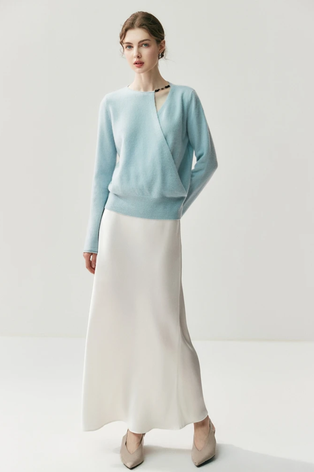Woman wearing a light blue sweater with a cut out on the shoulder paired with a white skirt on a plain background