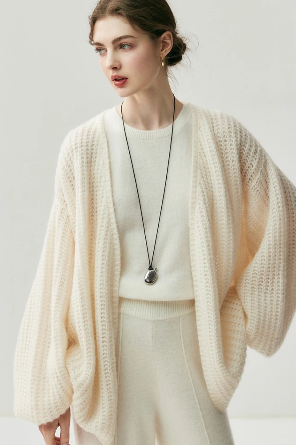 Woman wearing a cream-colored knit cardigan over a white top and pants on a light gray background