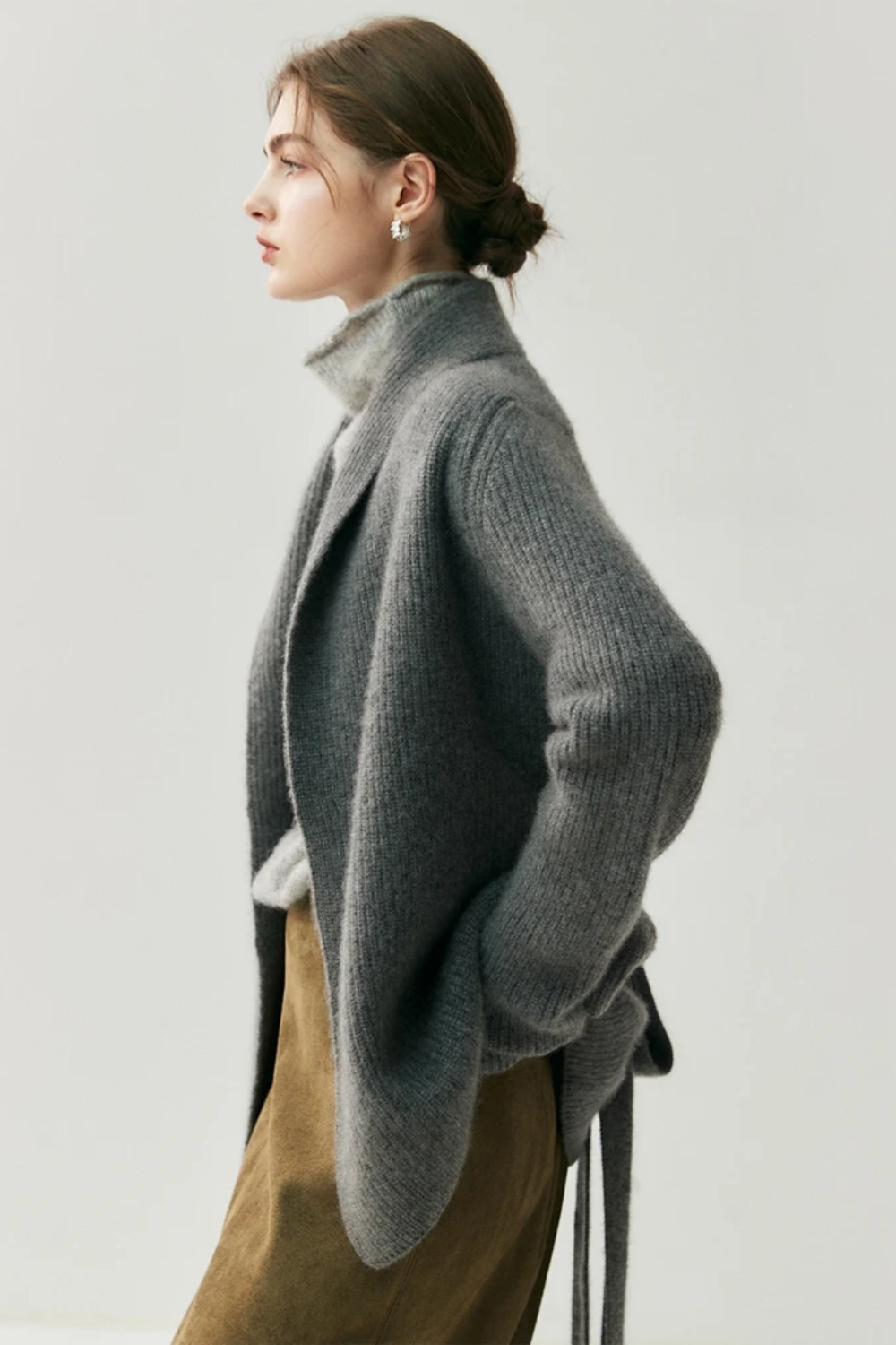 Woman wearing a gray cardigan over a turtleneck sweater facing the side with a plain background