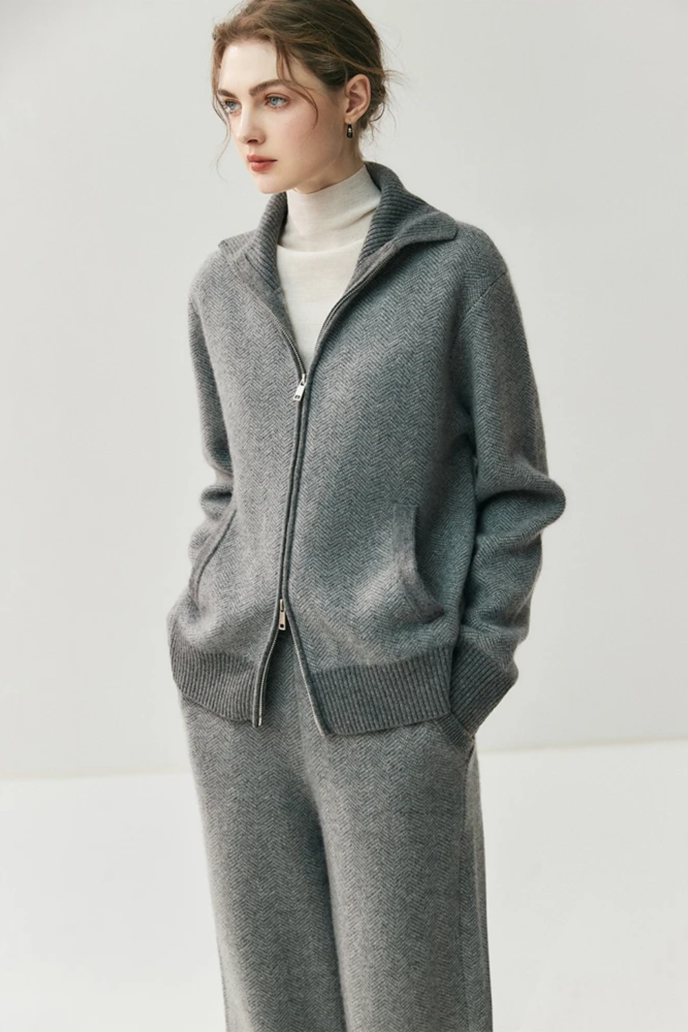 Woman wearing a gray zip-up sweater over a white turtleneck on a plain background