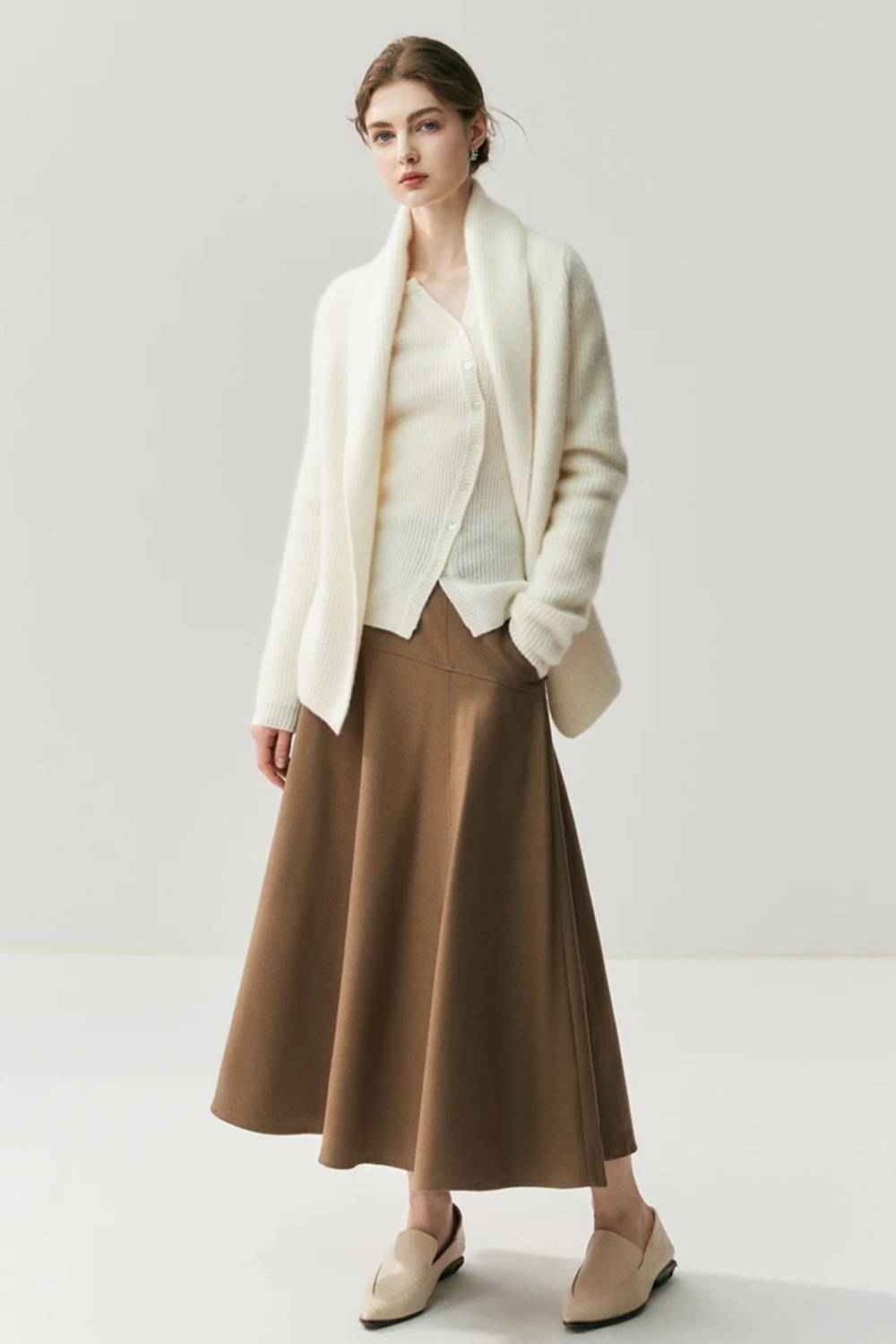 Woman wearing a white cardigan, beige blouse, brown skirt, and beige shoes on a white background