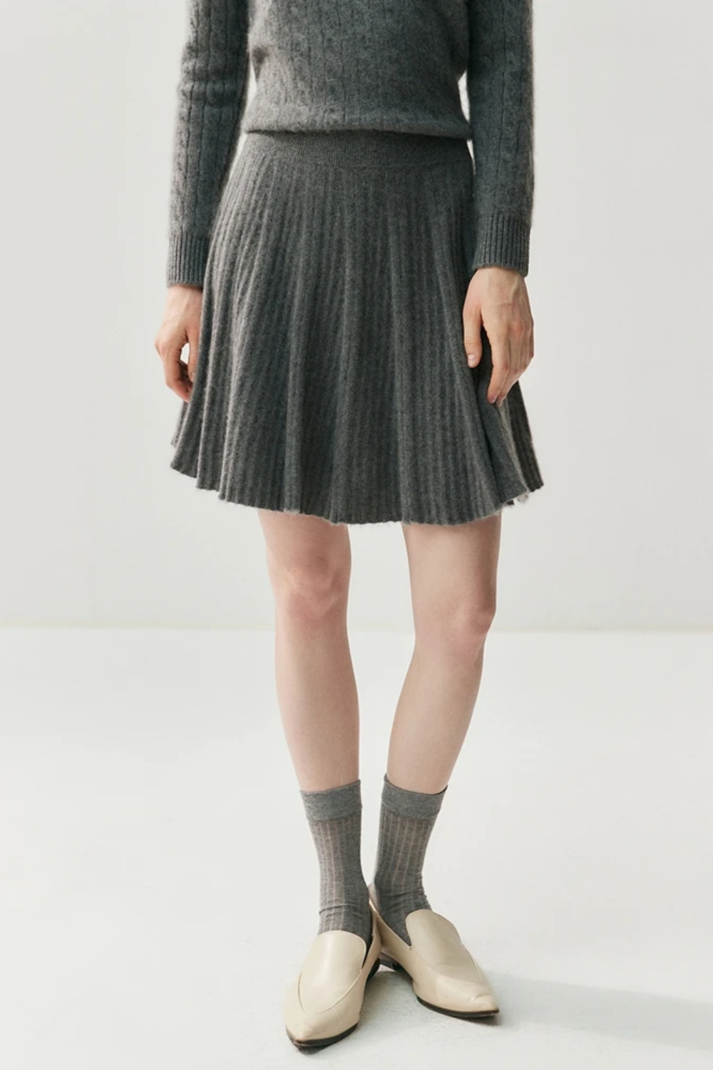 Person wearing a gray sweater, pleated skirt, and socks on a white background