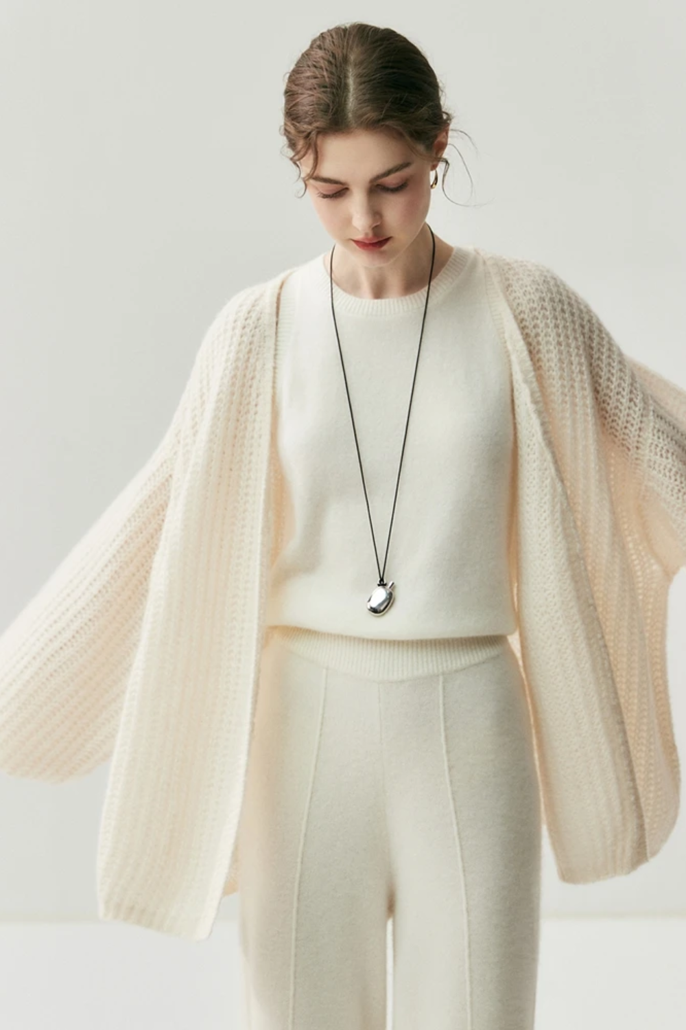 Woman wearing a cream-colored knit cardigan over a white top and pants on a light gray background