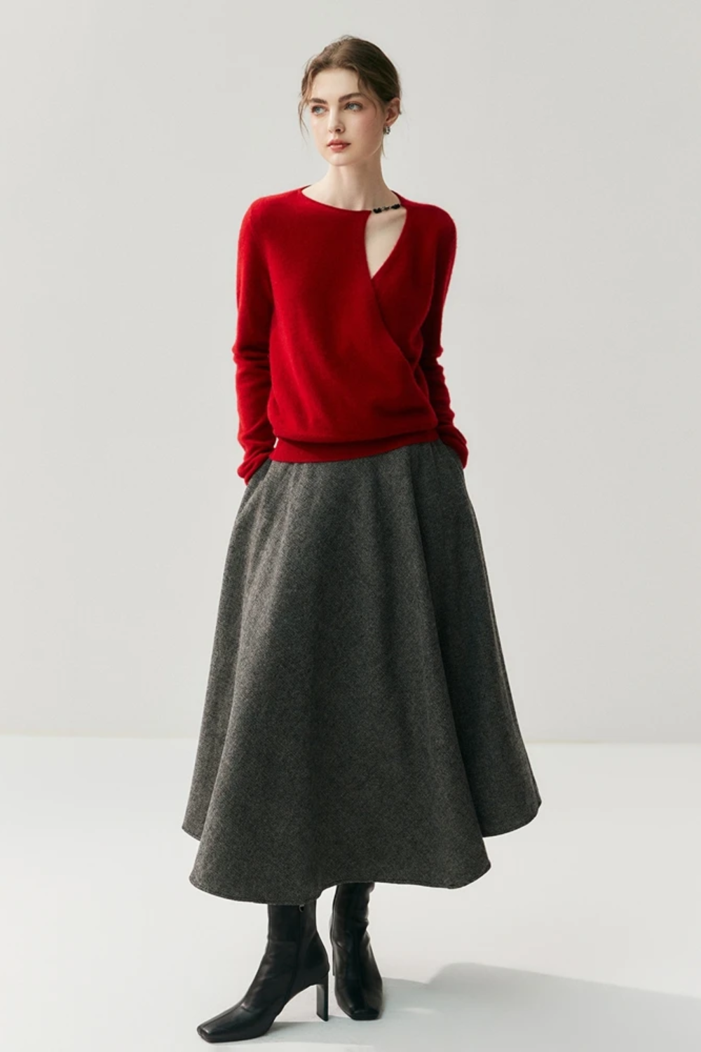 Woman wearing a red sweater with a cut out on the shoulder paired with a grey skirt on a plain background
