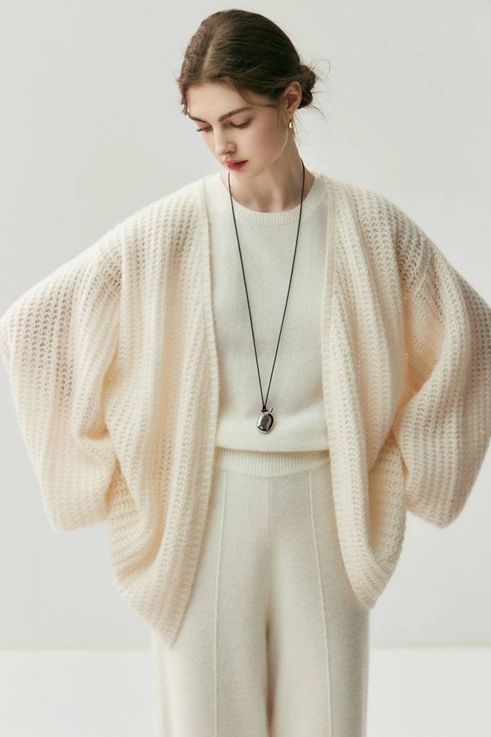 Woman wearing a cream-colored knit cardigan over a white top and pants on a light gray background