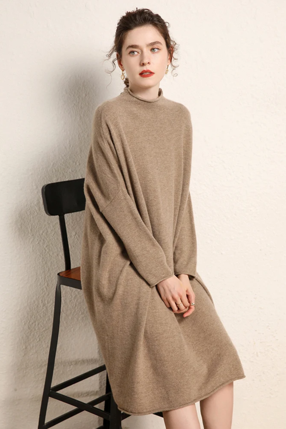 Woman wearing a camel sweater dress sitting on a black chair against a plain background