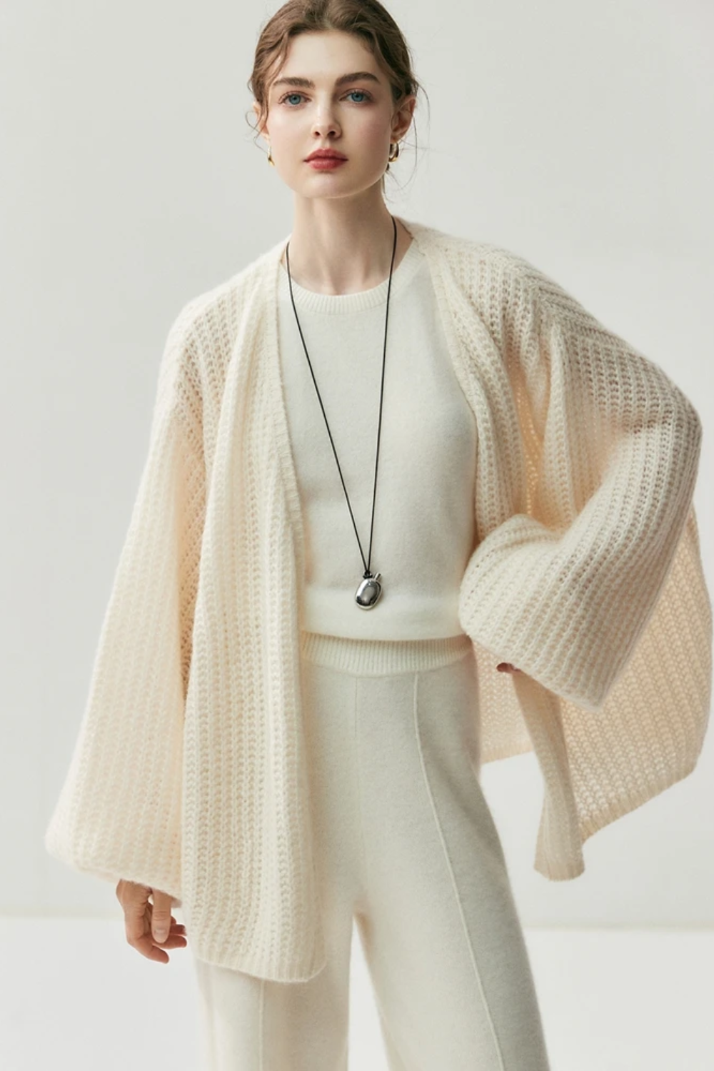 Woman wearing a cream-colored knit cardigan over a white top and pants on a light gray background