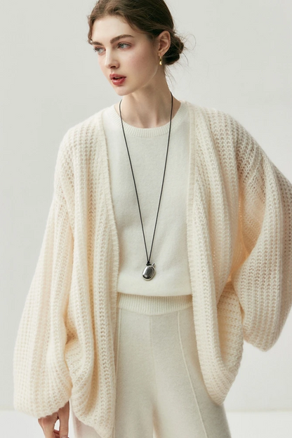 Woman wearing a cream-colored knit cardigan over a white top and pants on a light gray background