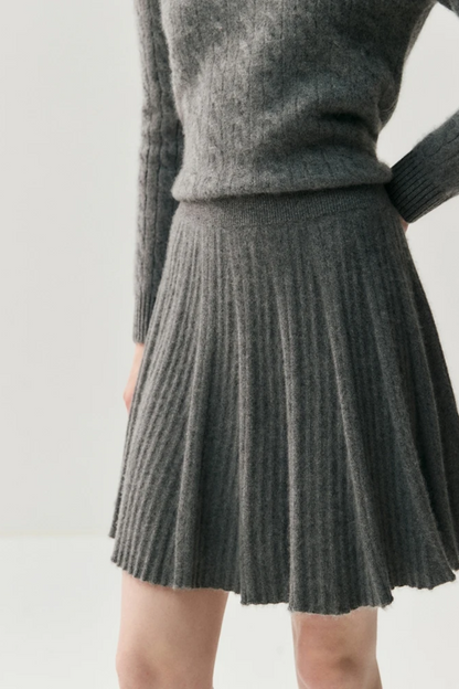 Person wearing a gray ribbed sweater and pleated skirt on a light background