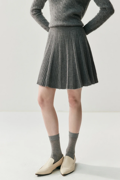 Person wearing a gray sweater, pleated skirt, and white shoes on a plain background