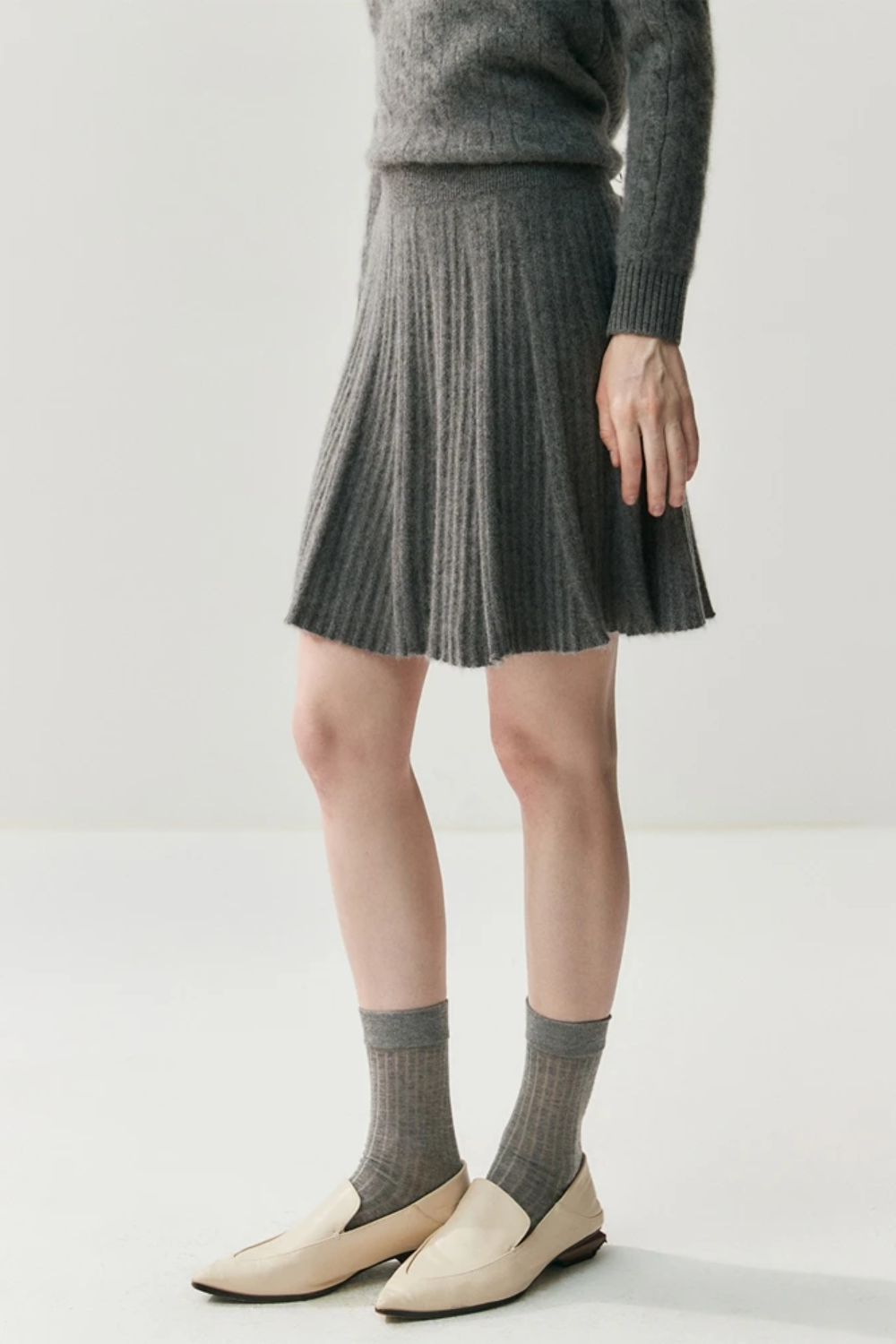 Person wearing a gray pleated skirt, gray sweater, and beige shoes on a white background