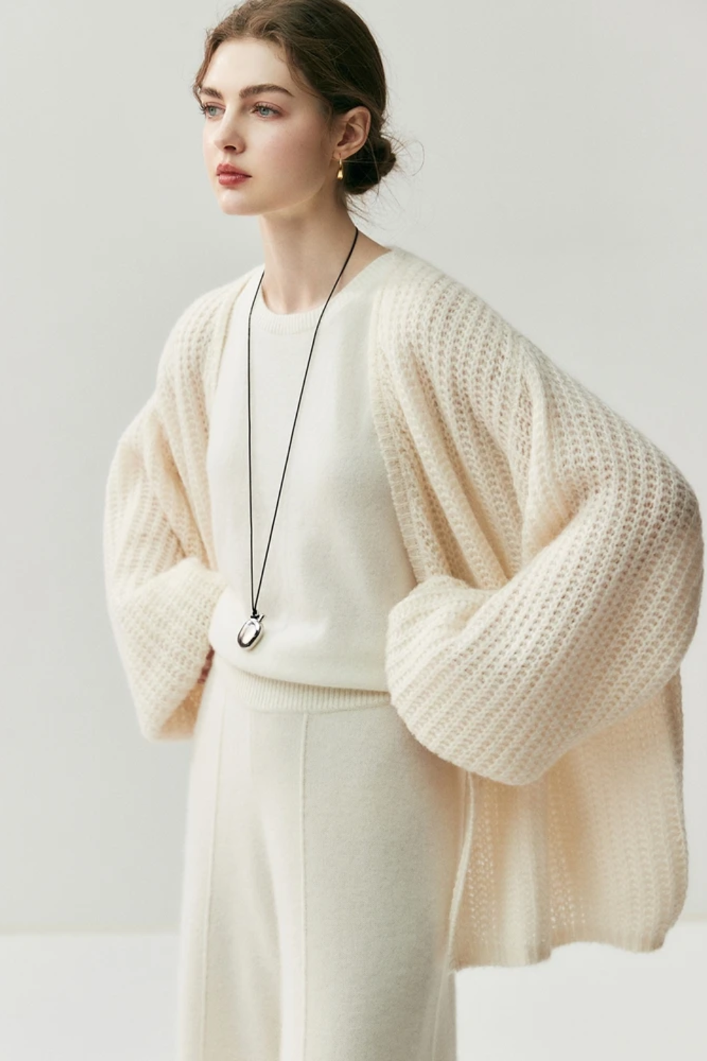 Woman wearing a cream-colored knit cardigan over a white top and pants on a light gray background